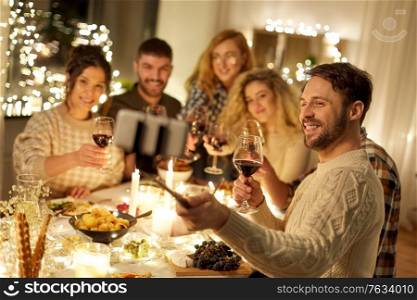 holidays, celebration and people concept - happy friends drinking non-alcoholic red wine and taking picture with smartphone on selfie stick at home christmas dinner party. friends taking selfie at christmas dinner party