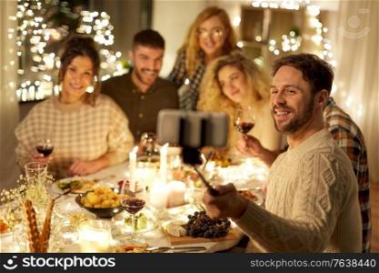holidays, celebration and people concept - happy friends drinking non-alcoholic red wine and taking picture with smartphone on selfie stick at home christmas dinner party. friends taking selfie at christmas dinner party