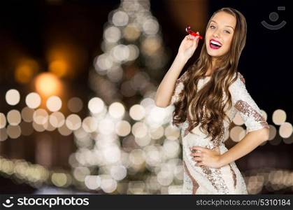 holidays, celebration and people concept - happy young woman or teen girl in fancy dress with sequins and party blower over christmas tree lights background. woman with party blower over christmas tree lights