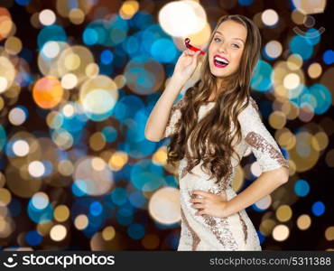 holidays, celebration and people concept - happy young woman or teen girl in fancy dress with sequins and party blower over festive lights background. happy woman with party blower over festive lights