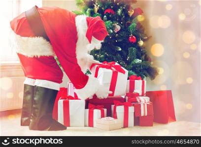 holidays, celebration and people concept - man in costume of santa claus putting present under christmas tree over lights