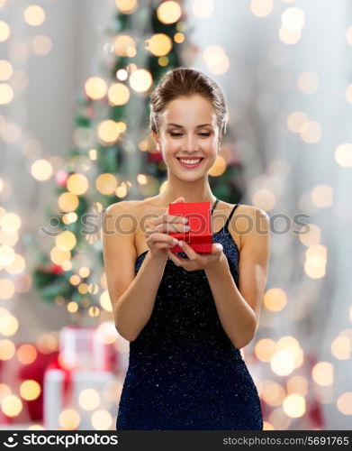 holidays, celebration and people concept - smiling woman in dress holding red gift box over christmas tree lights background