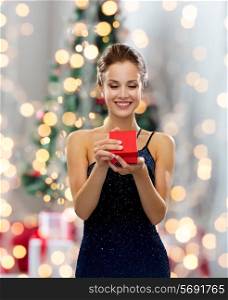 holidays, celebration and people concept - smiling woman in dress holding red gift box over christmas tree lights background
