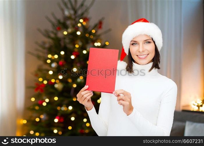 holidays, celebration and people concept - smiling woman in santa hat holding greeting card over christmas tree lights background. smiling woman with greeting card on christmas