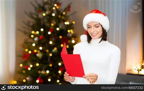 holidays, celebration and people concept - smiling woman in santa hat holding greeting card over christmas tree lights background. smiling woman with greeting card on christmas