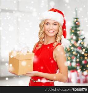 holidays, celebration and people concept - smiling woman in santa helper hat and red dress with gift box over living room and christmas tree background
