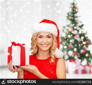 holidays, celebration and people concept - smiling woman in santa helper hat and red dress with gift box over living room and christmas tree background