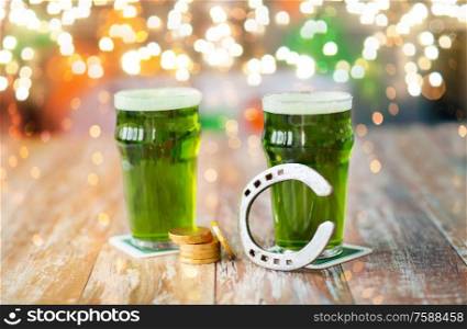 holidays, celebration and st patricks day concept - glasses of green beer, horseshoe and gold coins on table over festive lights. glasses of green beer, horseshoe and gold coins
