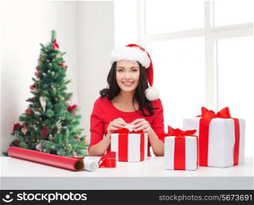 holidays, celebration, decoration and people concept - smiling woman in santa helper hat with decorating paper packing gift boxes over living room and christmas tree background
