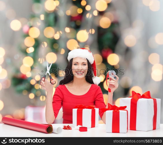 holidays, celebration, decoration and people concept - smiling woman in santa helper hat with scissors packing gift boxes over christmas tree and lights background