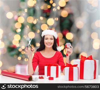 holidays, celebration, decoration and people concept - smiling woman in santa helper hat with scissors packing gift boxes over christmas tree and lights background