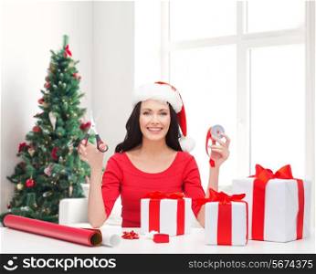 holidays, celebration, decoration and people concept - smiling woman in santa helper hat with scissors packing gift boxes over living room and christmas tree background