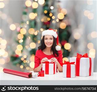 holidays, celebration, decoration and people concept - smiling woman in santa helper hat with decorating paper packing gift boxes over christmas tree lights background