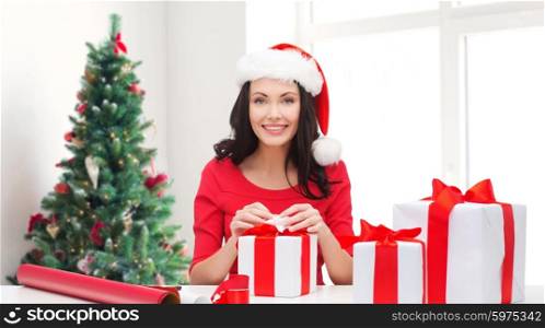 holidays, celebration, decoration and people concept - smiling woman in santa helper hat with decorating paper packing gift boxes over living room and christmas tree background