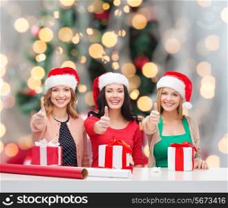 holidays, celebration, decoration and people concept - smiling women in santa helper hats with decorating paper and gift boxes showing thumbs up over christmas tree background