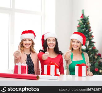 holidays, celebration, decoration and people concept - smiling women in santa helper hats with decorating paper and gift boxes showing thumbs up over living room and christmas tree background