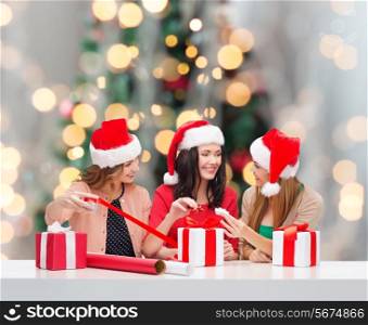 holidays, celebration, decoration and people concept - smiling women in santa helper hats with decorating paper and gift boxes over christmas tree background