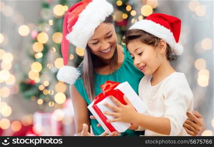 holidays, celebration, family and people concept - happy mother and little girl in santa helper hats with gift box over living room and christmas tree background
