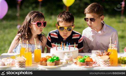holidays, childhood and celebration concept - happy kids blowing out candles on birthday cake sitting at table at summer garden party. happy kids with cake on birthday party at summer. happy kids with cake on birthday party at summer