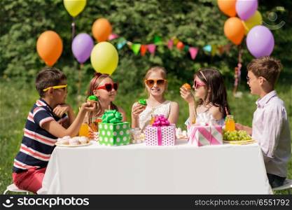 holidays, childhood and celebration concept - happy kids in sunglasses sitting at table on birthday party at summer garden and eating cupcakes. kids eating cupcakes on birthday party at summer. kids eating cupcakes on birthday party at summer