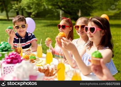 holidays, childhood and celebration concept - happy kids in sunglasses sitting at table on birthday party at summer garden and eating cupcakes. kids eating cupcakes on birthday party in summer