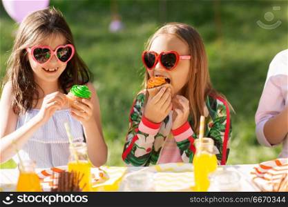 holidays, childhood and celebration concept - happy kids in sunglasses sitting at table on birthday party at summer garden and eating cupcakes. kids eating cupcakes on birthday party at summer. kids eating cupcakes on birthday party at summer