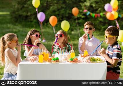 holidays, childhood and celebration concept - happy kids with cake blowing party horns and having fun at summer birthday. happy kids blowing party horns at summer birthday. happy kids blowing party horns at summer birthday