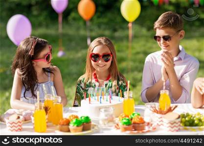 holidays, childhood and celebration concept - happy kids with candles on birthday cake sitting at table at summer garden party. happy kids with cake on birthday party at summer. happy kids with cake on birthday party at summer