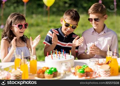 holidays, childhood and celebration concept - happy kids with candles on cake sitting at table at summer garden party and applauding to birthday boy. happy kids with cake on birthday party at summer. happy kids with cake on birthday party at summer
