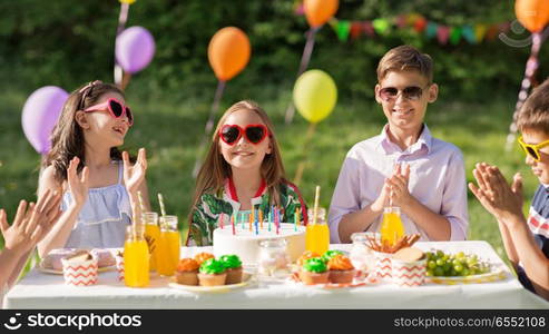 holidays, childhood and celebration concept - happy kids with candles on cake sitting at table at summer garden party and applauding to birthday girl. happy kids with cake on birthday party at summer. happy kids with cake on birthday party at summer