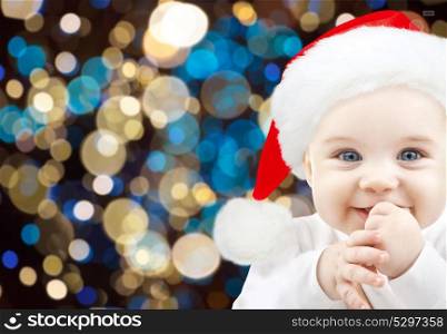 holidays, childhood and people concept - happy baby in santa hat over lights background. happy baby in santa hat over christmas lights