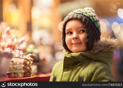 holidays, childhood and people concept - happy little boy at christmas market candy shop in winter evening. happy little boy at christmas market shop