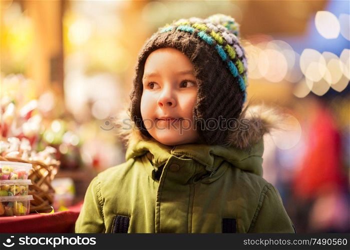 holidays, childhood and people concept - happy little boy at christmas market candy shop in winter evening. happy little boy at christmas market in winter