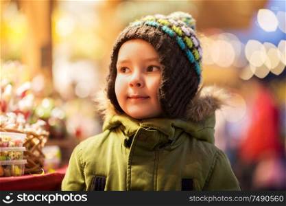 holidays, childhood and people concept - happy little boy at christmas market candy shop in winter evening. happy little boy at christmas market in winter