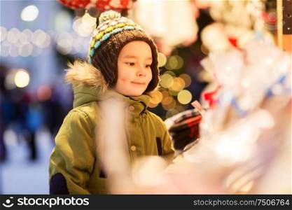 holidays, childhood and people concept - happy little boy at christmas market stall in winter evening. happy little boy at christmas market in winter