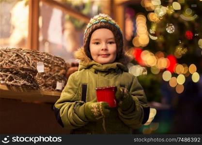 holidays, childhood and people concept - happy little boy with cup of tea at christmas market in winter evening. happy boy with cup of tea at christmas market