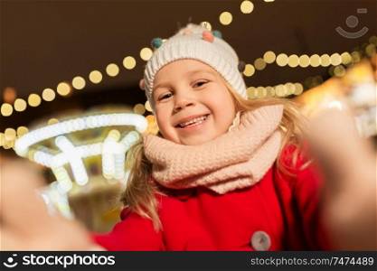 holidays, childhood and people concept - happy little girl at christmas market taking selfie in winter evening. little girl taking selfie at christmas market
