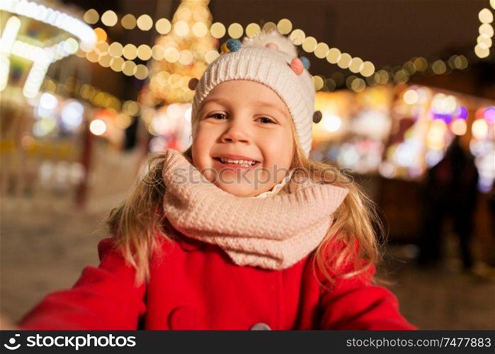 holidays, childhood and people concept - happy little girl at christmas market taking selfie in winter evening. little girl taking selfie at christmas market