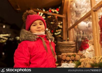 holidays, childhood and people concept - happy little girl at market shop window in winter. girl at christmas market
