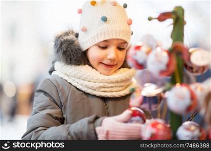 holidays, childhood and people concept - happy little girl choosing christmas balls at market in winter. little girl choosing christmas balls at market