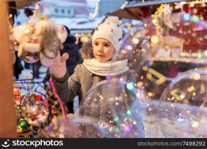 holidays, childhood and people concept - happy little girl choosing christmas balls at market in winter. little girl choosing christmas balls at market