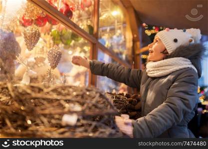 holidays, childhood and people concept - happy little girl choosing christmas decorations at market shop window in winter. girl choosing christmas decorations at market