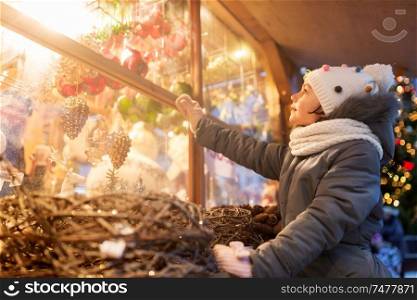 holidays, childhood and people concept - happy little girl choosing christmas decorations at market shop window in winter. girl choosing christmas decorations at market