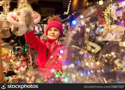 holidays, childhood and people concept - happy little girl choosing christmas decorations at market in winter. girl choosing christmas decorations at market