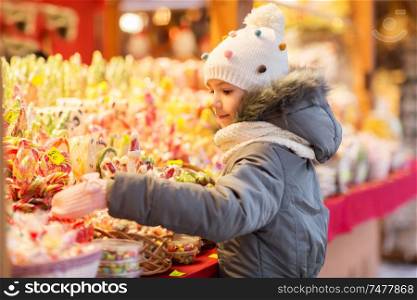 holidays, childhood and people concept - happy little girl choosing sweets at christmas market in winter evening. little girl choosing sweets at christmas market