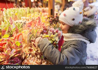 holidays, childhood and people concept - happy little girl choosing sweets at christmas market in winter evening. little girl choosing sweets at christmas market