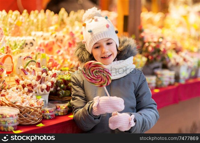 holidays, childhood and people concept - happy little girl with big lollipop at christmas market candy shop in evening. girl with lollipop at christmas market candy shop