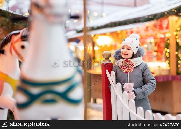 holidays, childhood and people concept - happy little girl with big lollipop at christmas market. girl with lollipop at christmas market