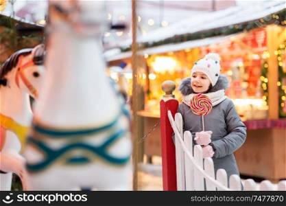 holidays, childhood and people concept - happy little girl with big lollipop at christmas market. girl with lollipop at christmas market