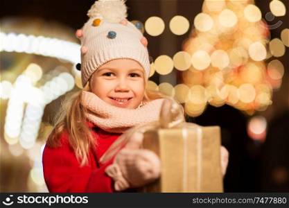 holidays, childhood and people concept - happy little girl with gift box at christmas market in winter evening. happy girl with gift box at christmas market